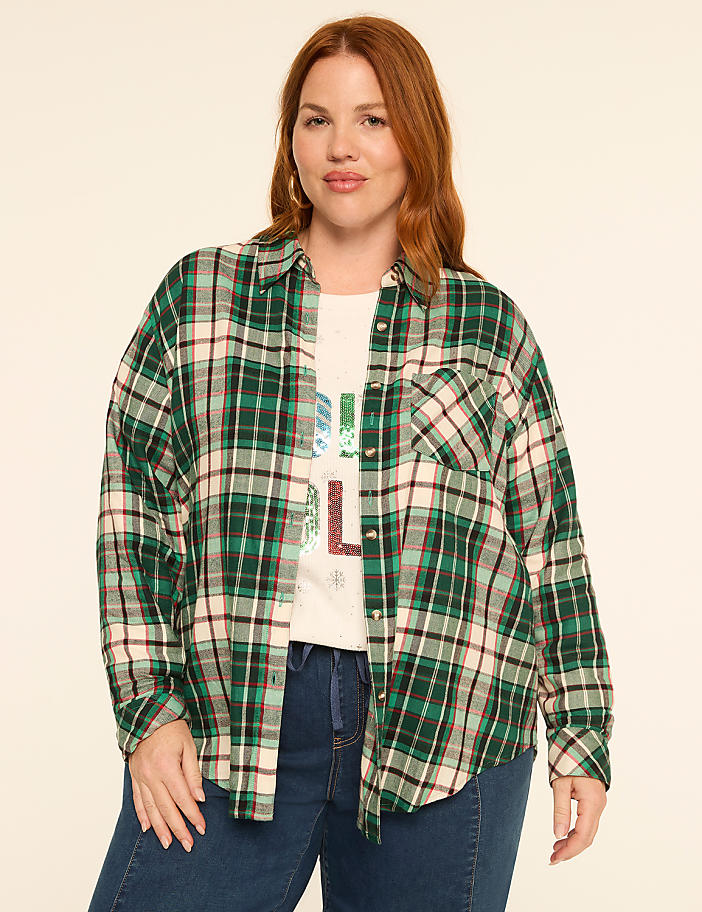A woman with red hair and a plaid shirt stands against a plain, light-colored background.