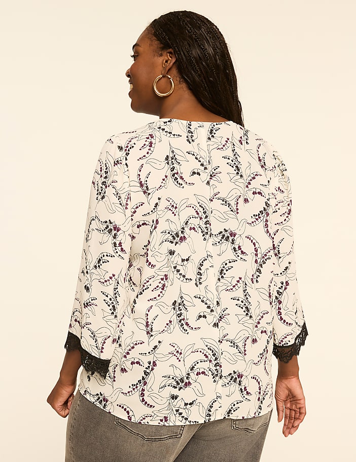 A woman in a floral print blouse and gray jeans stands against a plain, light-colored background.