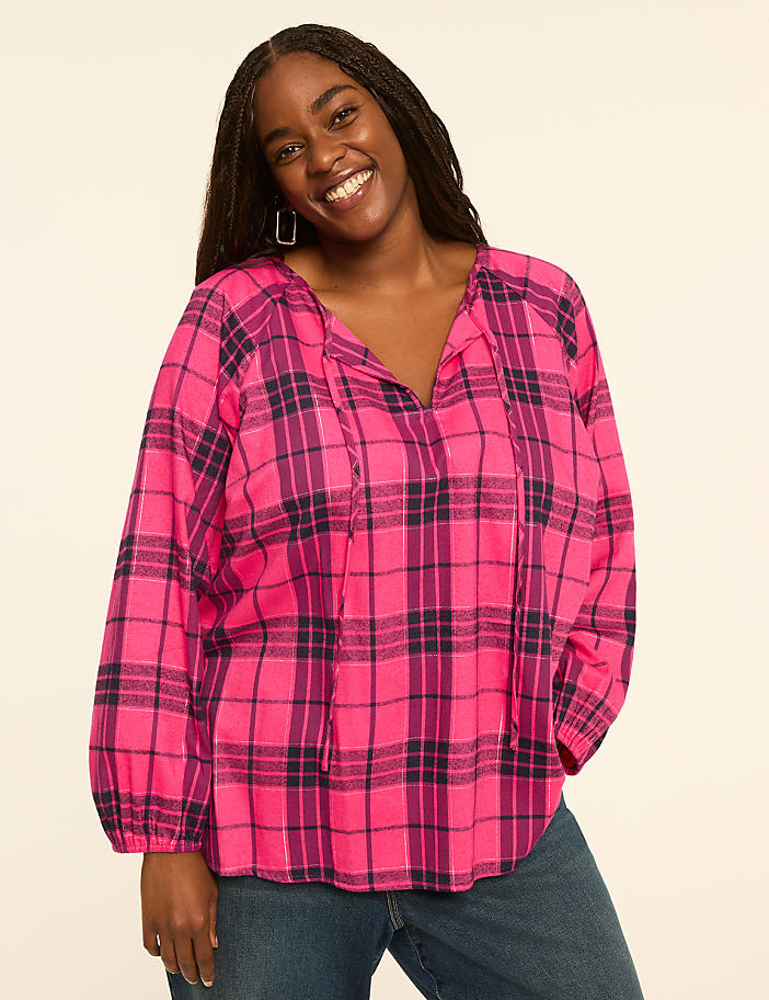 A smiling woman in a pink and purple plaid blouse stands against a plain, light-colored background.