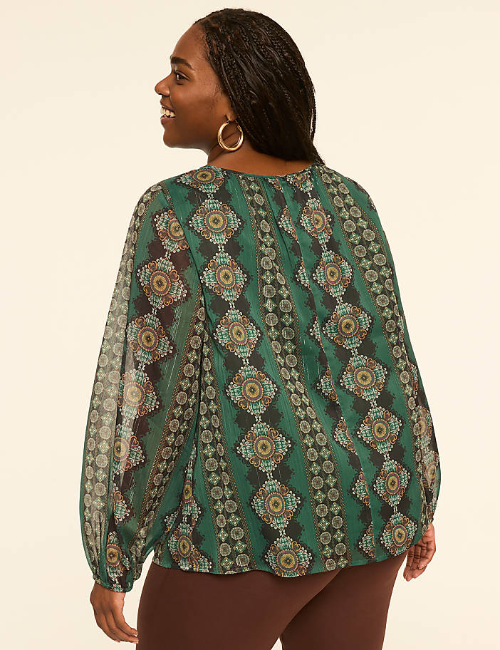 A woman in a patterned green blouse stands against a plain, light-colored background.