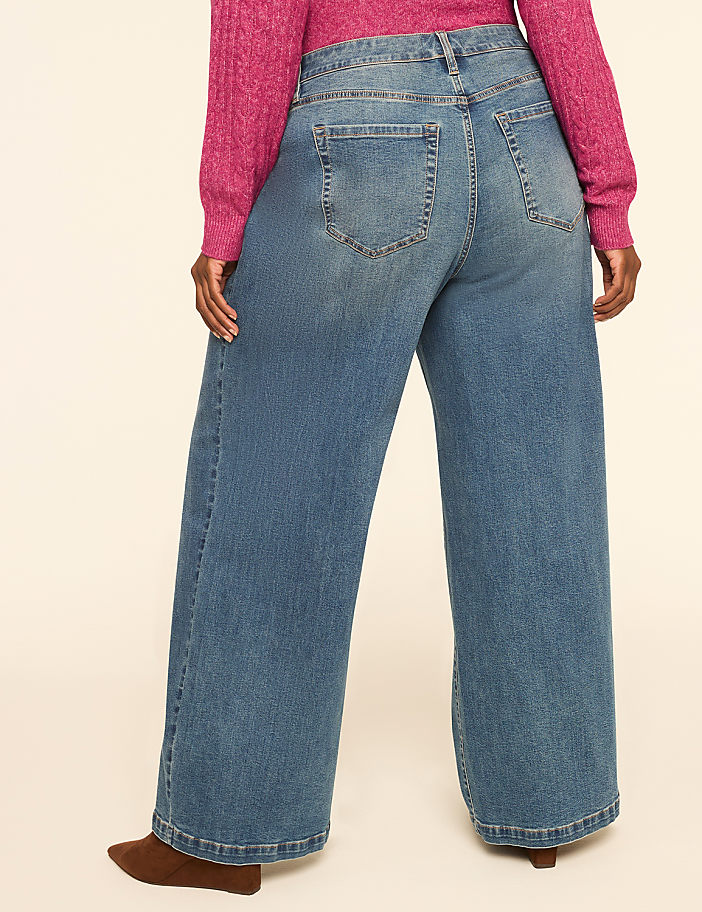 A person wearing wide-leg jeans and a pink sweater stands against a plain, light-colored background.