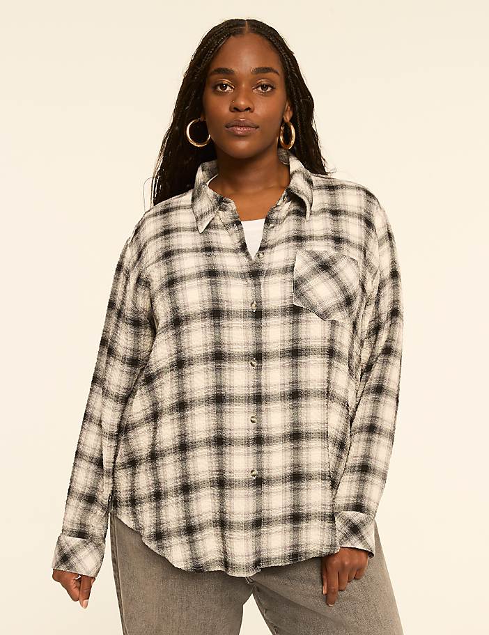 A woman with dark hair and hoop earrings poses in a cream and black plaid shirt and grey jeans against a plain, light-colored background.
