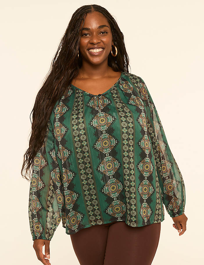 A smiling woman with long dark hair wears a flowing green patterned blouse and brown leggings against a plain cream background.