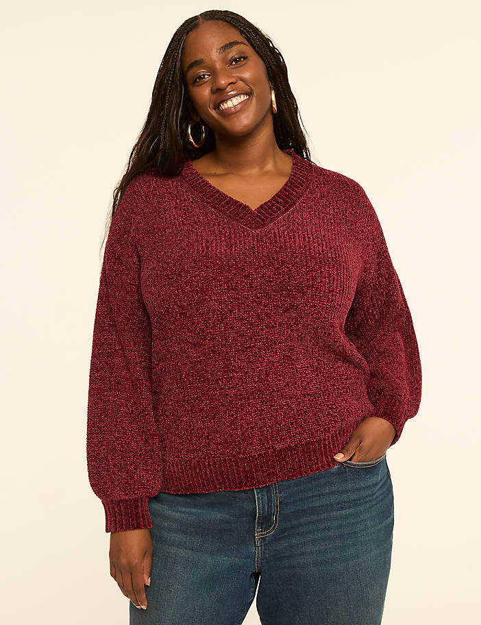 A smiling woman with dark braided hair, wearing a burgundy v-neck sweater and blue jeans, stands against a plain cream background.