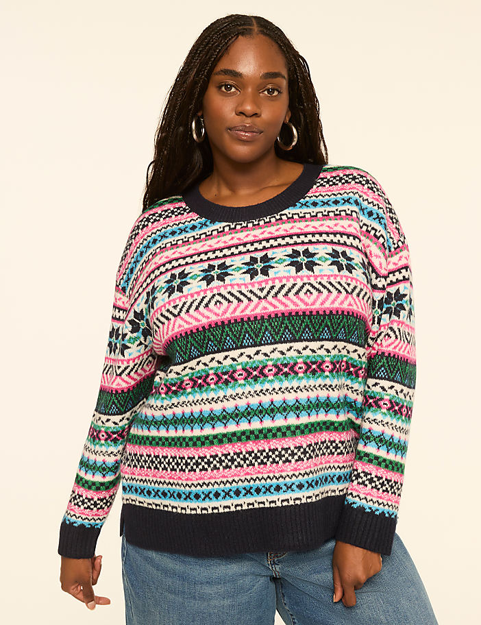 A woman with braided hair and hoop earrings poses in a colorful, patterned sweater and blue jeans against a plain, light-colored background.
