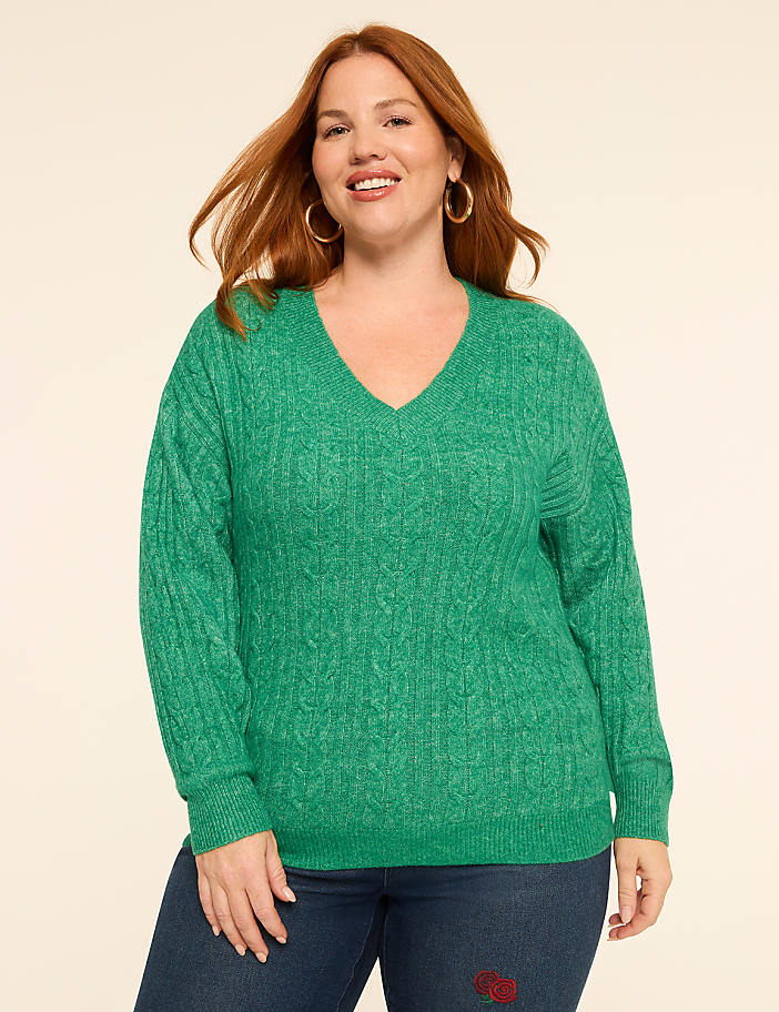 A woman with red hair smiles in a green cable-knit sweater and blue jeans against a plain, light-colored background.