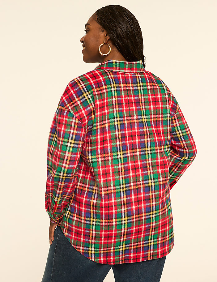 A woman in a red, green, and yellow plaid shirt stands against a plain, light-colored background.