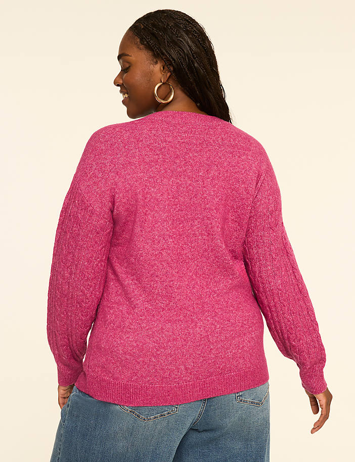 A woman in a pink sweater and jeans stands against a plain, light-colored background.