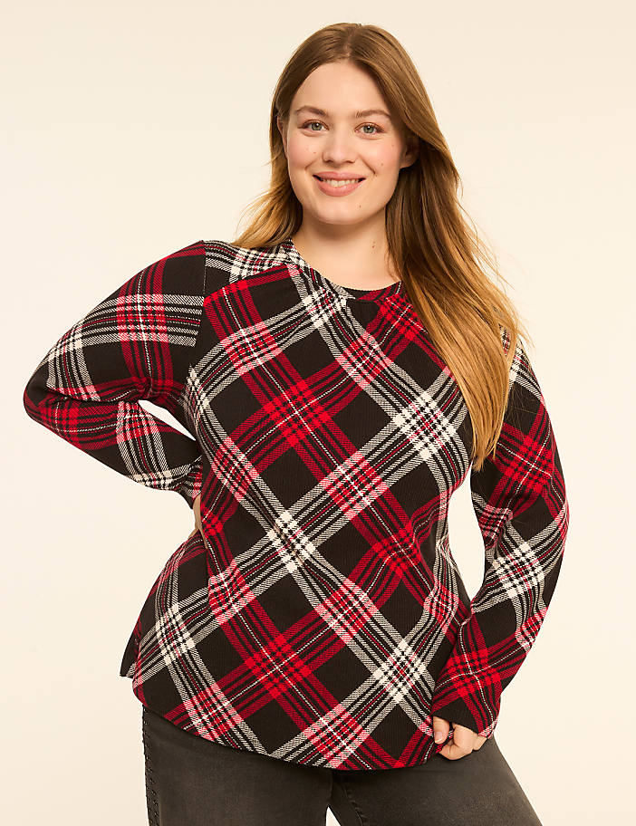 A smiling woman in a red, black, and white plaid shirt stands against a plain, light-colored background.