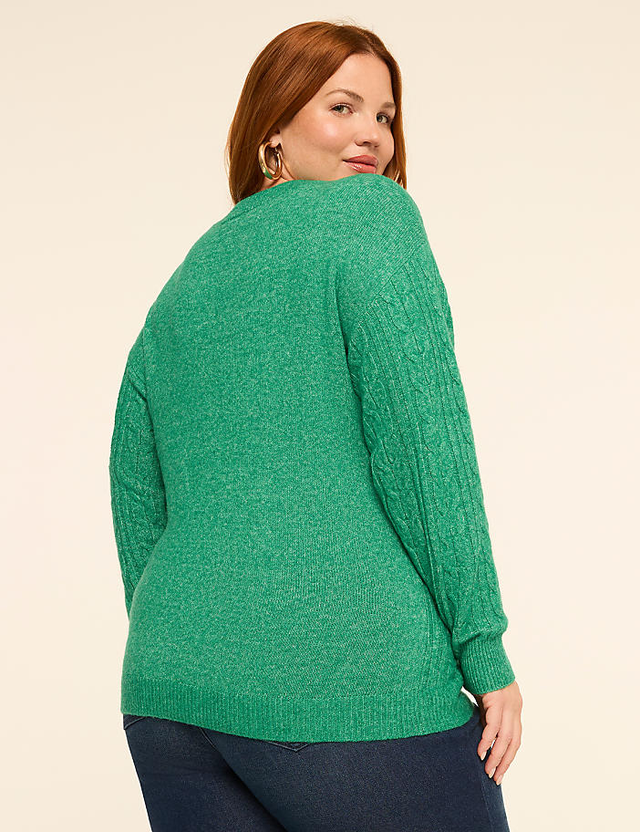 A woman with auburn hair wearing a green cable-knit sweater and dark jeans stands against a plain, light-colored background.