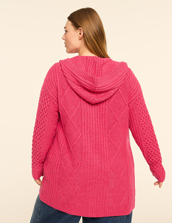 A woman in a pink hooded cardigan stands against a plain, light-colored background.