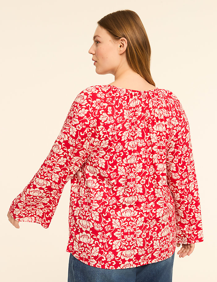 A woman in a red floral blouse stands against a plain, light-colored background.