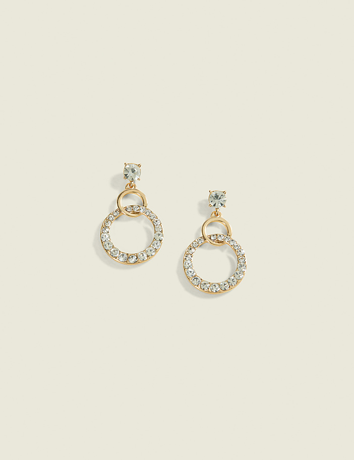A pair of gold hoop earrings encrusted with clear rhinestones dangle against a soft, muted beige background.