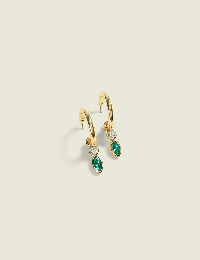 A pair of gold hoop earrings with emerald and diamond pendants hang against a plain, light-colored background.
