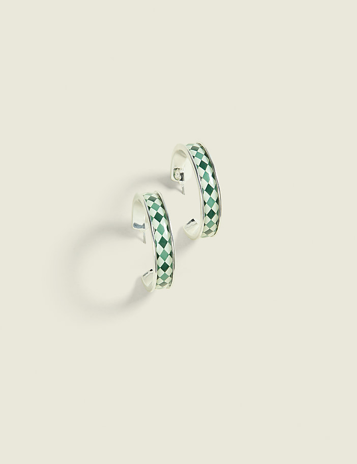 A pair of silver hoop earrings with a green diamond pattern rests on a pale, textured background.