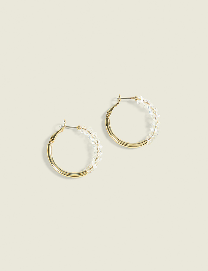 A pair of gold hoop earrings adorned with small clear beads are displayed against a soft, pale yellow background.