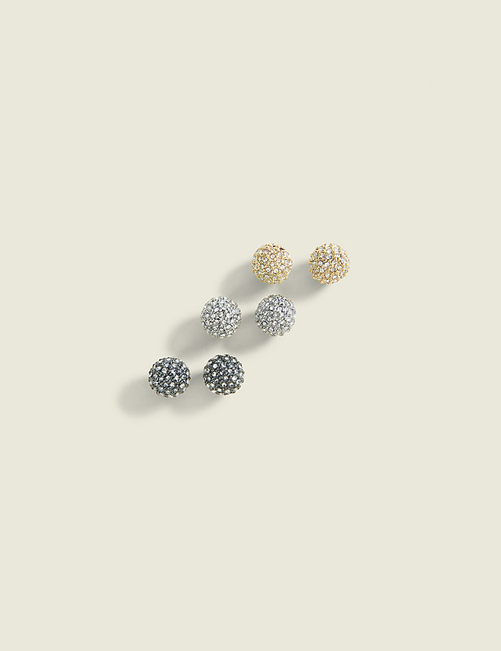 Three pairs of sparkling, spherical stud earrings in gold, silver, and black are arranged diagonally on a plain, light beige background.