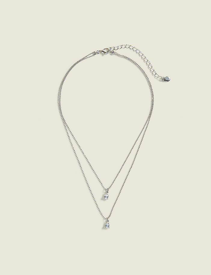 A delicate silver layered necklace with two small diamond pendants hangs against a plain, light beige background.
