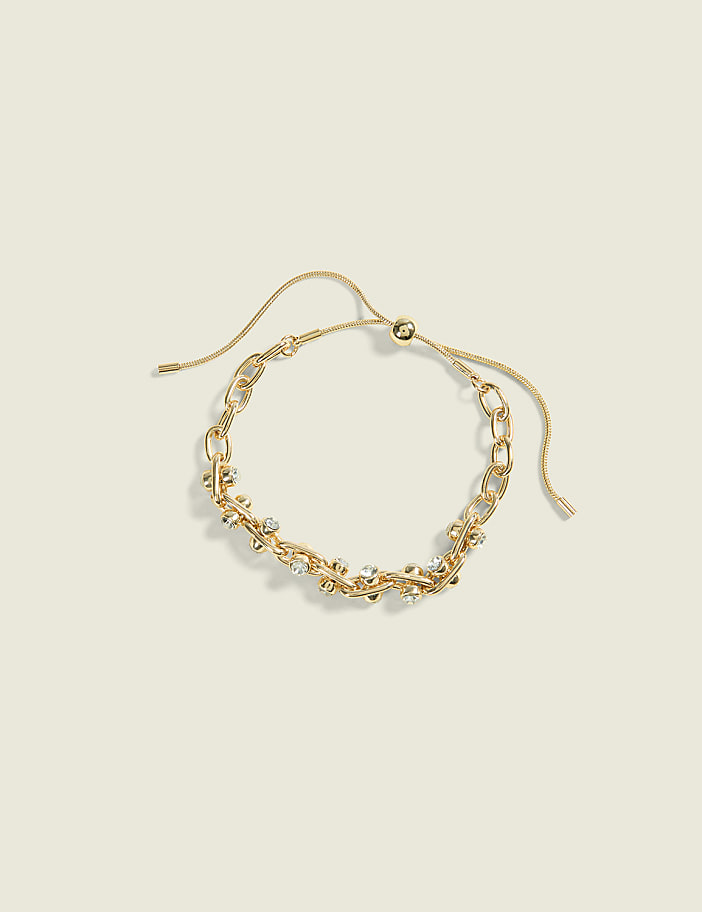 A delicate gold chain bracelet with sparkling stones rests on a soft, muted yellow background.