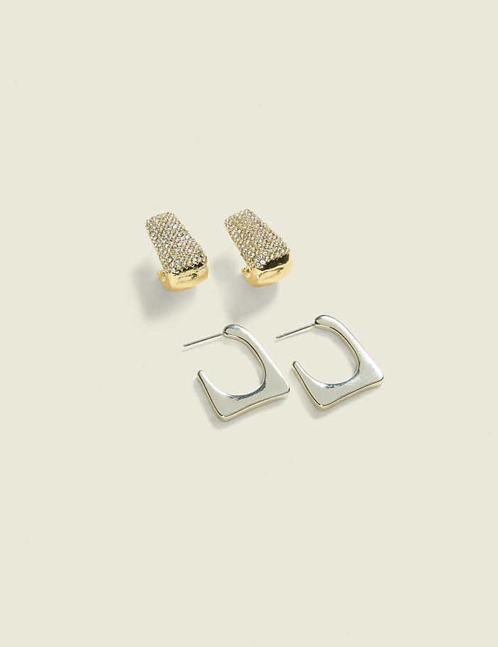 Two pairs of earrings, one gold and jeweled, the other silver and geometric, rest on a pale, textured background.