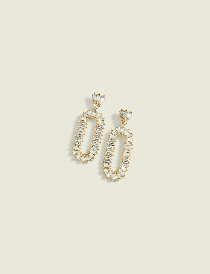 A pair of gold oval-shaped earrings with baguette-cut crystals dangle against a pale yellow background.