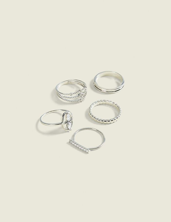 Five silver rings, some with intricate designs and others plain, are arranged on a pale, textured background.