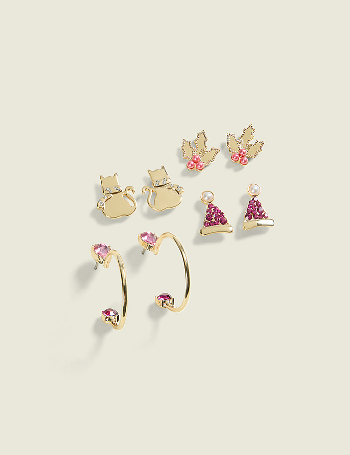 Gold earrings in the foreground feature pink gems and a hoop design, while the background displays a collection of festive stud earrings shaped like cats, holly leaves, and Santa hats.