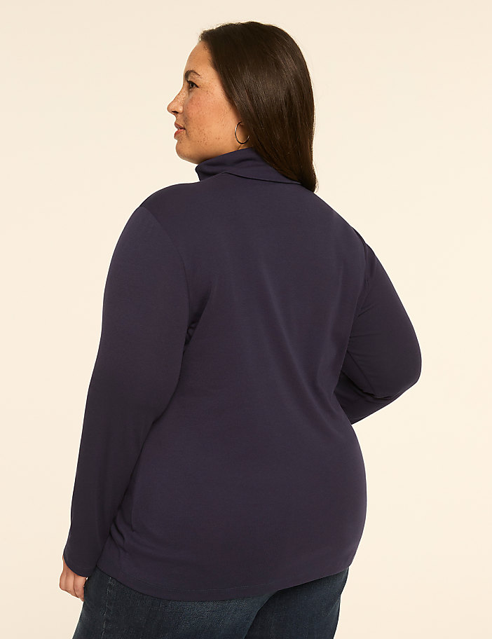 A woman in a navy turtleneck and jeans stands against a plain, light-colored background.