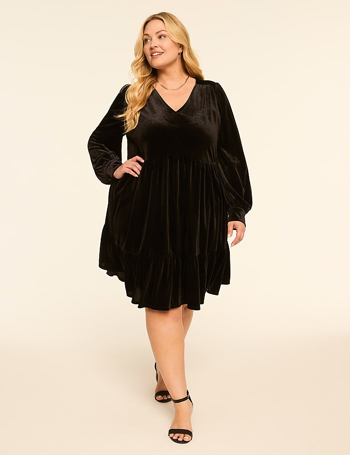 A woman in a black velvet dress stands against a plain, light-colored background.