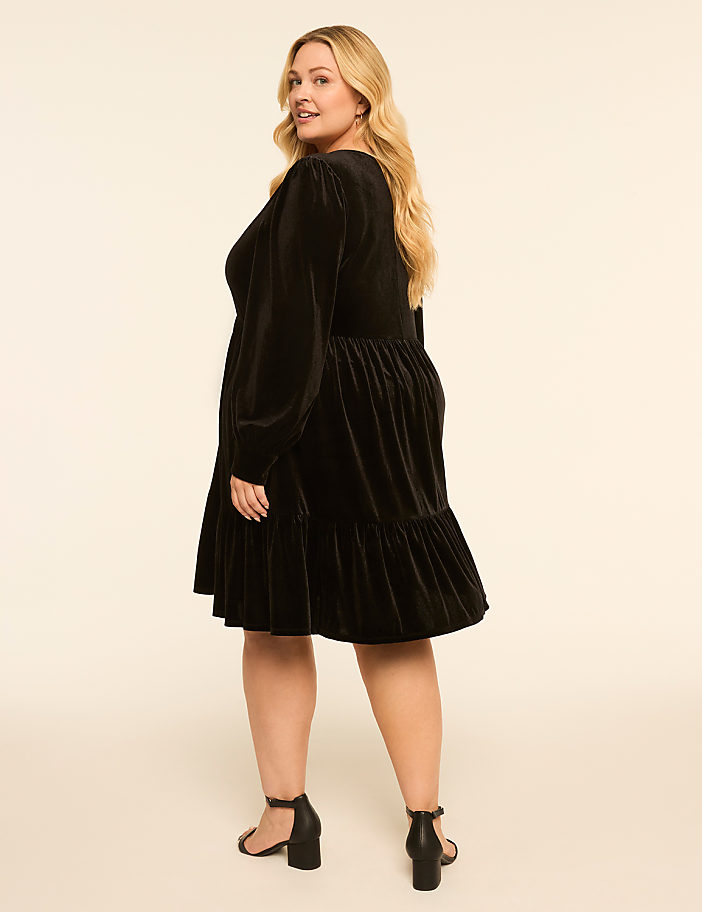 A blonde woman in a black velvet dress stands against a plain, light-colored background.