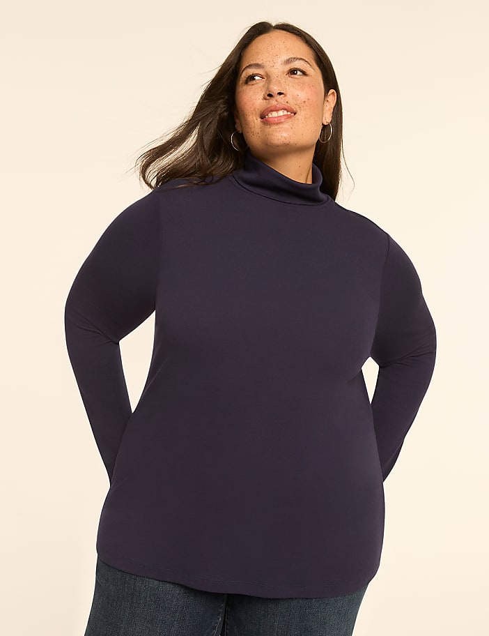 A woman in a navy turtleneck and jeans stands against a plain, light-colored background.