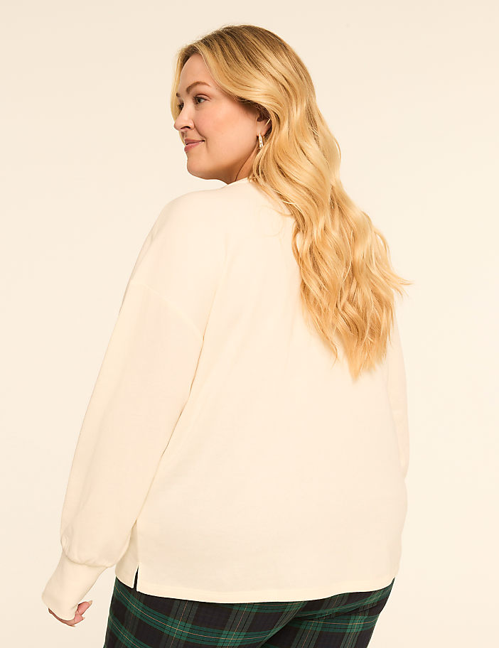 A blonde woman in a cream-colored sweatshirt and plaid pants stands against a plain, light-colored background.