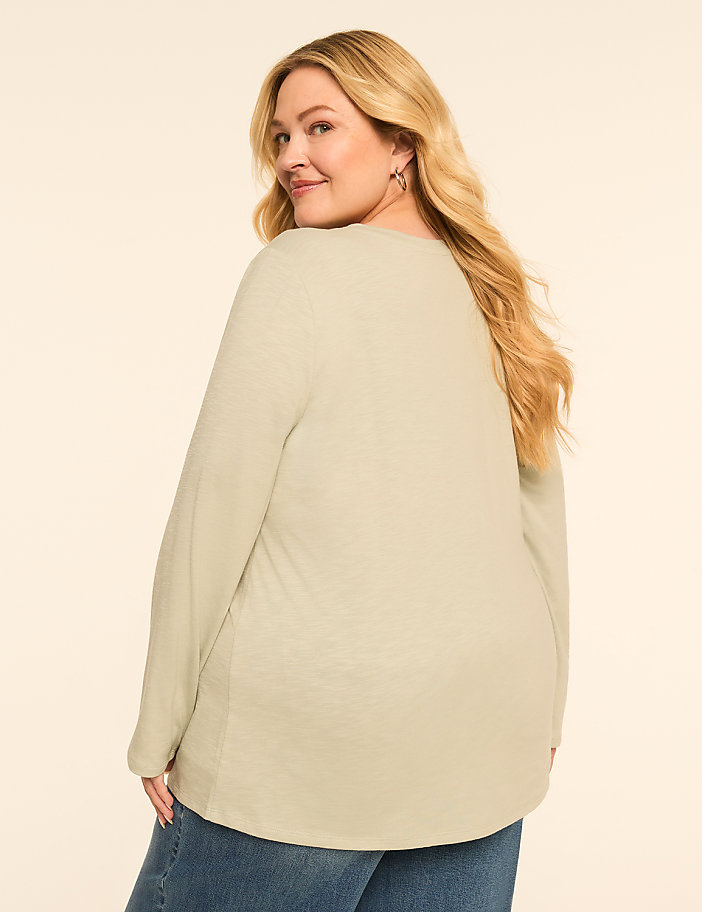 A woman in a light green long-sleeved shirt and blue jeans smiles over her shoulder against a plain, light-colored background.