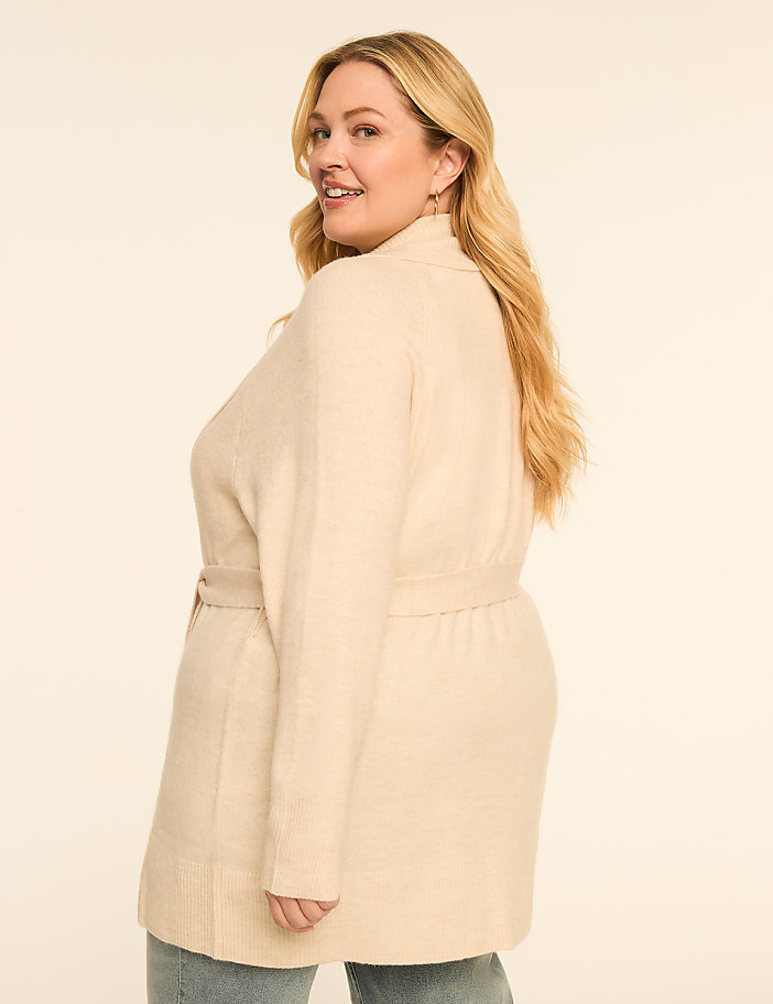 A woman in a cream-colored cardigan and jeans smiles over her shoulder against a plain, light background.