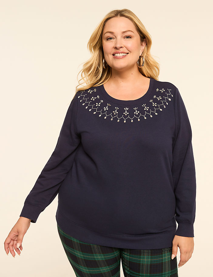 A smiling woman in a navy sweatshirt with a jeweled neckline and plaid pants stands against a plain, light background.
