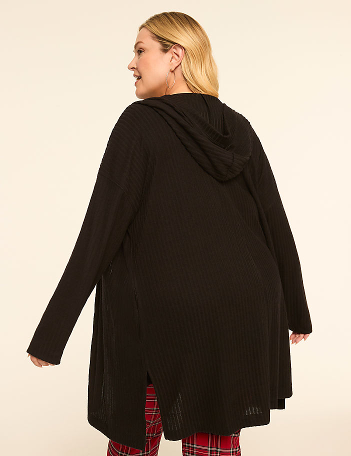 A woman in a black hooded cardigan stands against a plain, light-colored background, her plaid pajama bottoms visible at the hem.