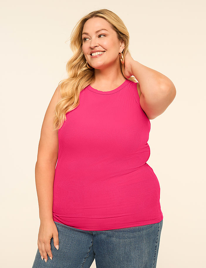A smiling woman with blonde hair wears a bright pink ribbed tank top and blue jeans against a plain, light-colored background.