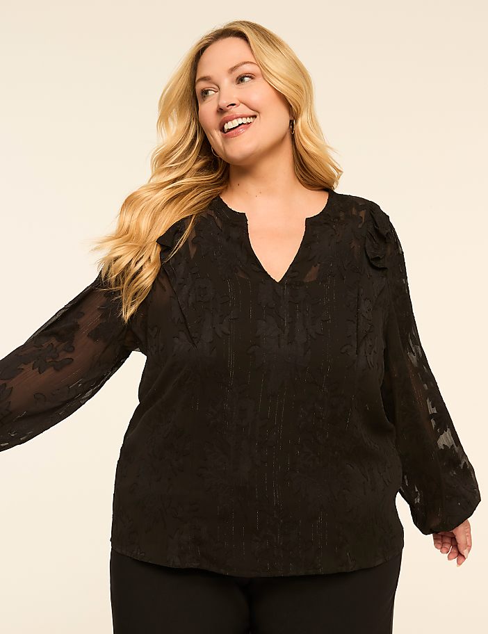 A smiling woman with blonde hair wears a black floral-patterned blouse with sheer sleeves, set against a plain, light-colored background.