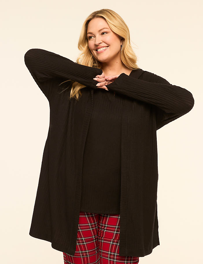 A smiling woman in a black cardigan and plaid pants stands against a plain, light-colored background.