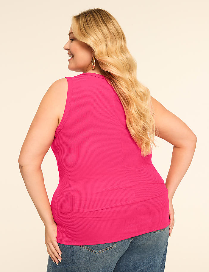 A woman in a bright pink ribbed tank top and blue jeans smiles over her shoulder against a plain, light-colored background.