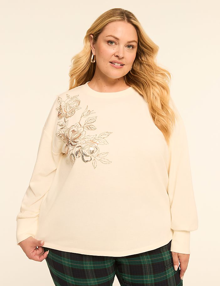 A woman with blonde hair and a cream-colored sweatshirt with floral embellishments stands against a plain, light-colored background, wearing plaid pants.
