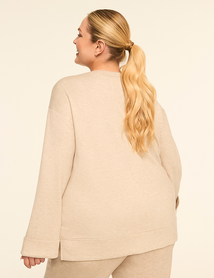 A woman with blonde hair in a ponytail, wearing a beige sweatshirt and pants, stands against a plain beige background.