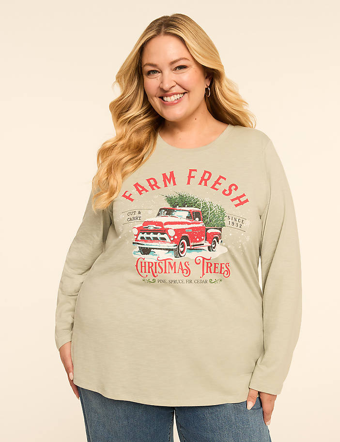 A smiling woman in a light green long-sleeved shirt with a vintage red truck graphic featuring Christmas trees stands against a plain background.