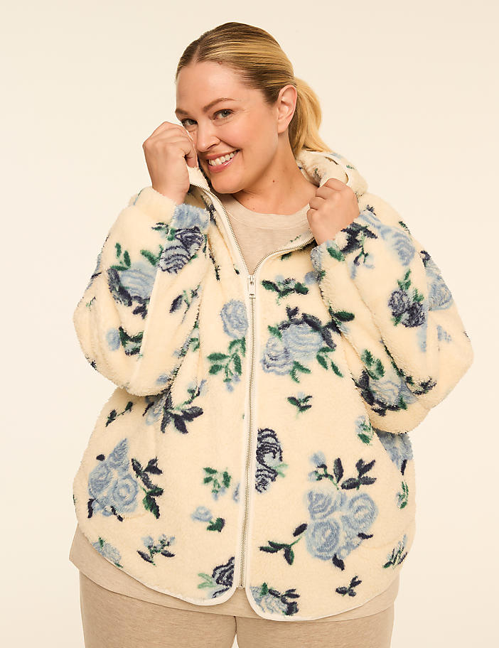 A smiling woman in a cream-colored, floral-patterned fleece jacket stands against a plain, light-colored background.