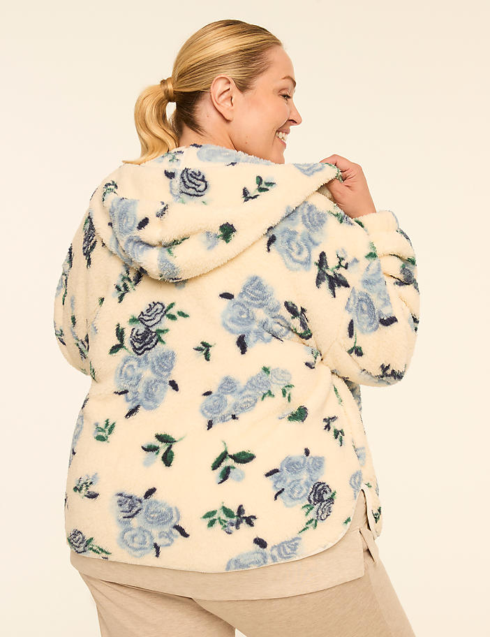 A woman in a cream-colored fleece hoodie with a blue floral pattern smiles over her shoulder against a plain, light-colored background.