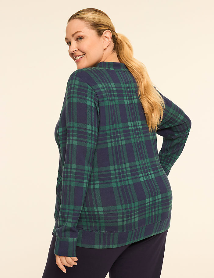 A woman in a green and navy plaid sweater and dark pants smiles over her shoulder against a plain, light background.