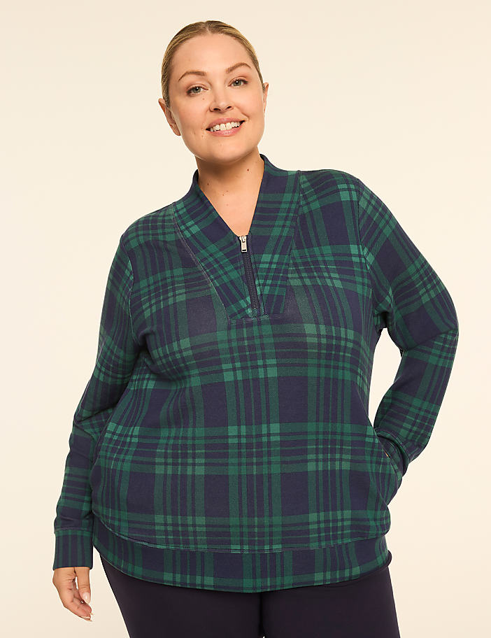 A smiling woman in a green and navy plaid quarter-zip sweatshirt stands against a plain, light-colored background.
