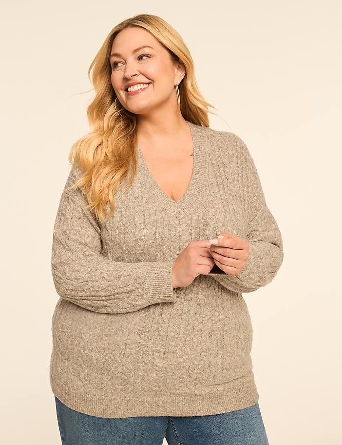 A smiling woman in a cable-knit sweater and jeans stands against a plain, light-colored background.