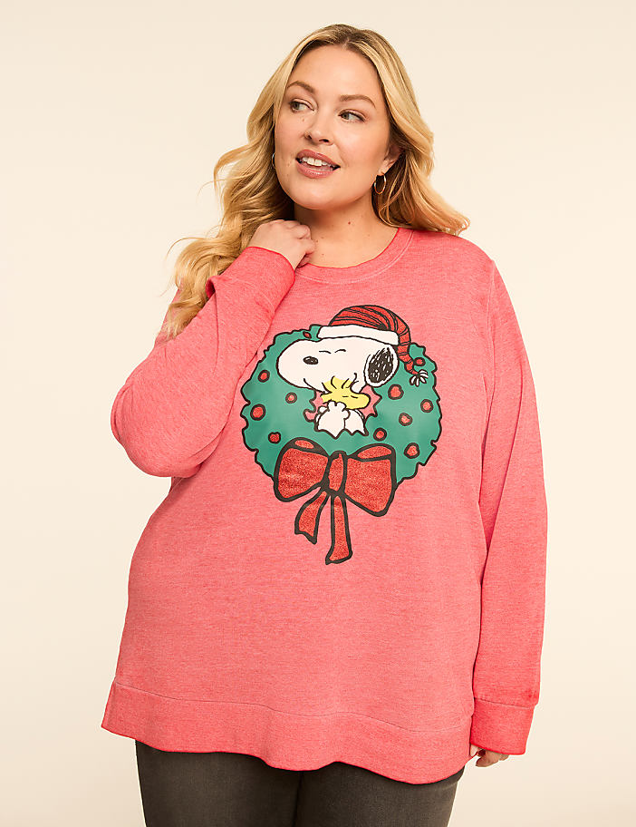 A woman in a coral sweatshirt with a Snoopy Christmas wreath design stands against a plain, light-colored background.