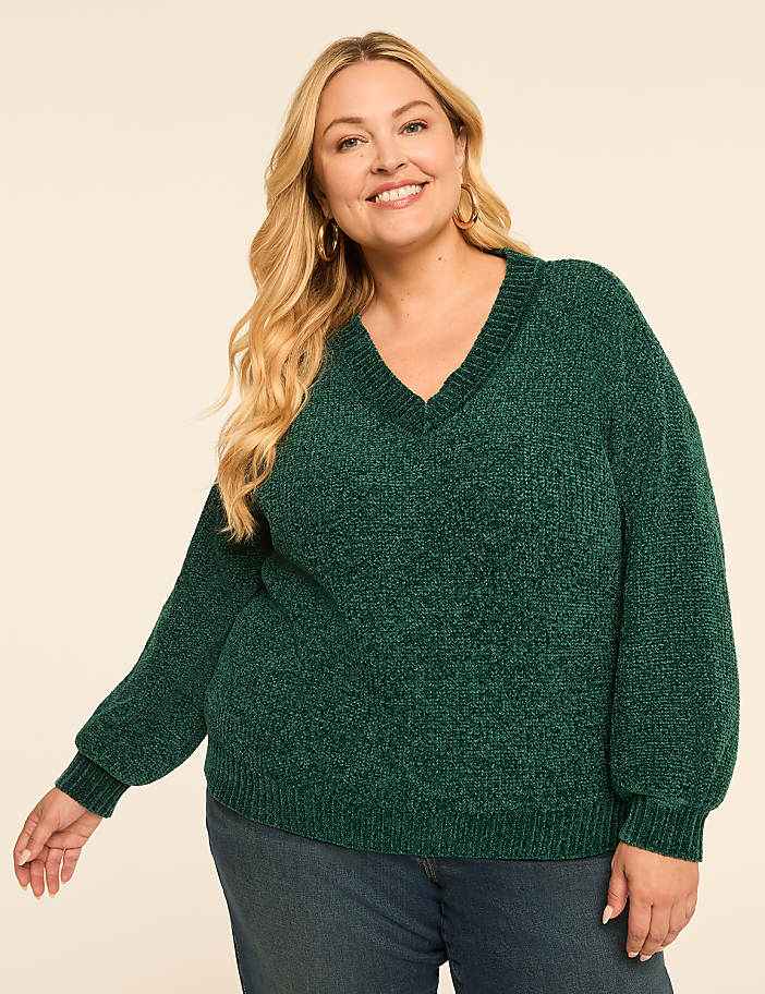 A smiling woman with blonde hair wears a green v-neck sweater and blue jeans against a plain, light-colored background.
